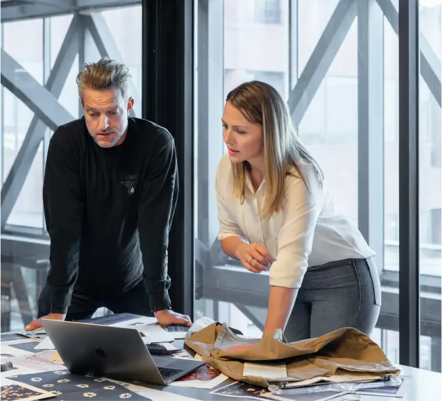 Two business professionals discussing ideas and reviewing documents on a desk with a laptop in a modern office setting.